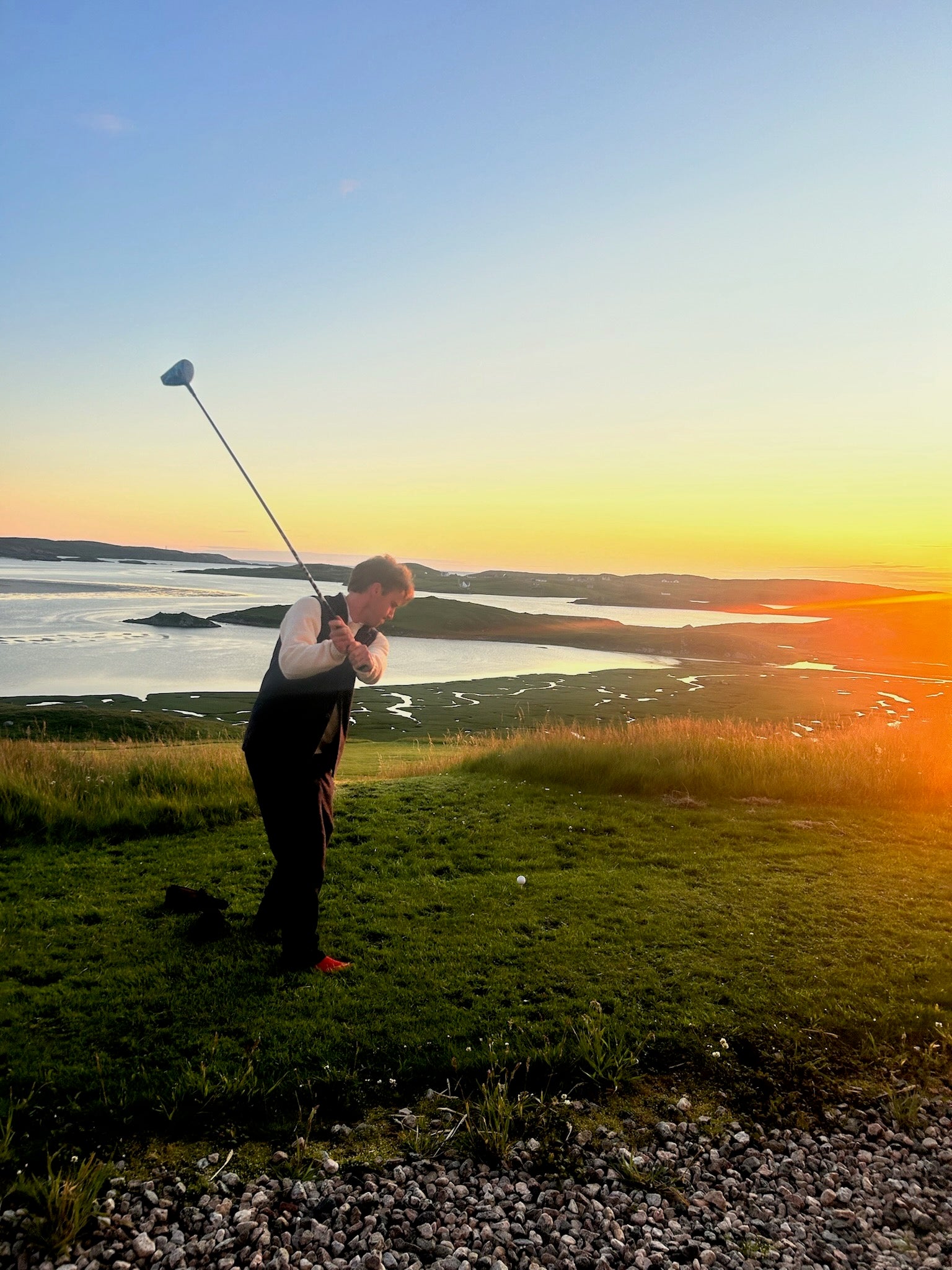 Man teeing off in front of Uig Lodge with a stunning sunset over Uig Bay in the background